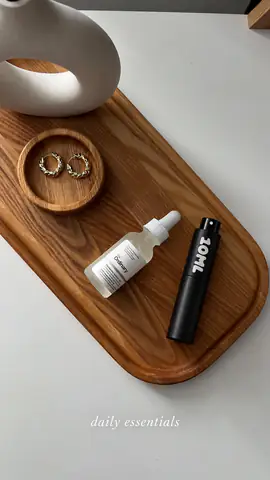 Černý rozprašovač 10ML na dřevěném podnosu se zlatými náušnicemi a sérem, s popiskem „daily essentials“